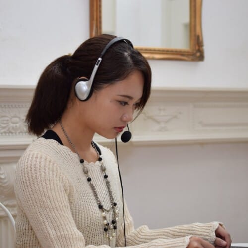 Woman wearing a headset with microphone, looking at a laptop on a desk in a bright room with a white ornate fireplace behind her