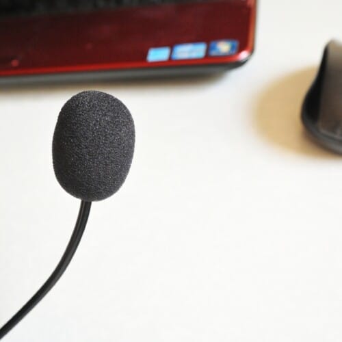 Close-up of a black foam-covered headset microphone on a stand with a laptop in the blurred background.