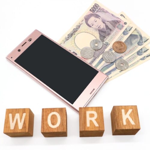 Pink smartphone rests on Japanese yen banknotes and coins, with wooden blocks spelling WORK in front.