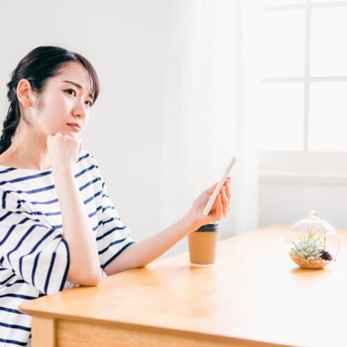 Woman in a striped shirt sits at a wooden table, looking at her smartphone with a thoughtful expression, in a bright room with a plant and coffee cup nearby