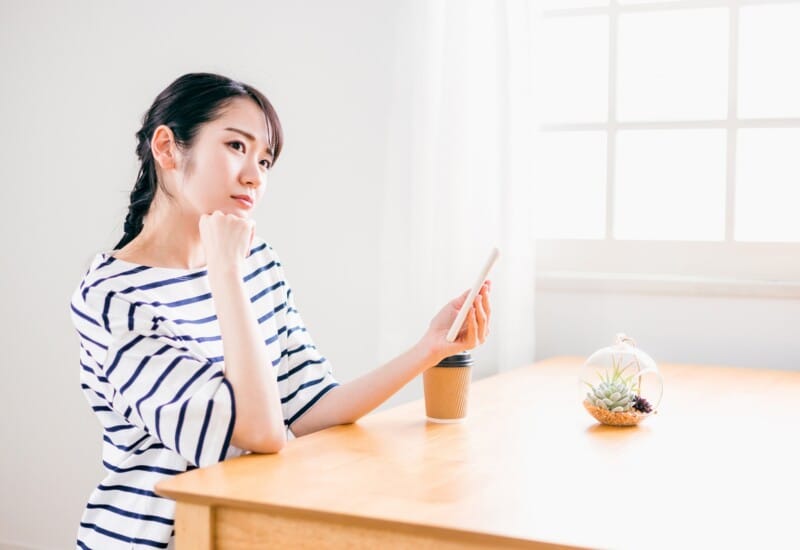 Woman in a striped shirt sits at a wooden table, looking at her smartphone with a thoughtful expression, in a bright room with a plant and coffee cup nearby