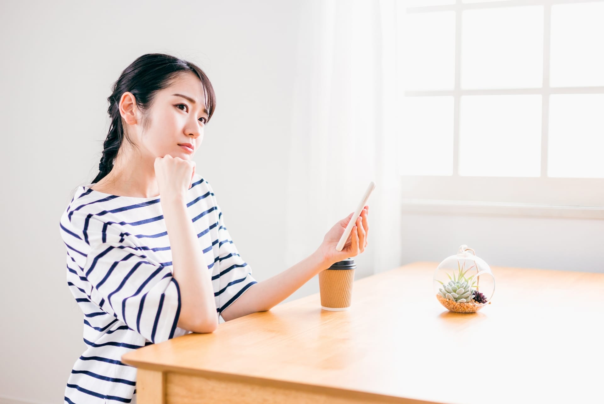 Woman in a striped shirt sits at a wooden table, looking at her smartphone with a thoughtful expression, in a bright room with a plant and coffee cup nearby
