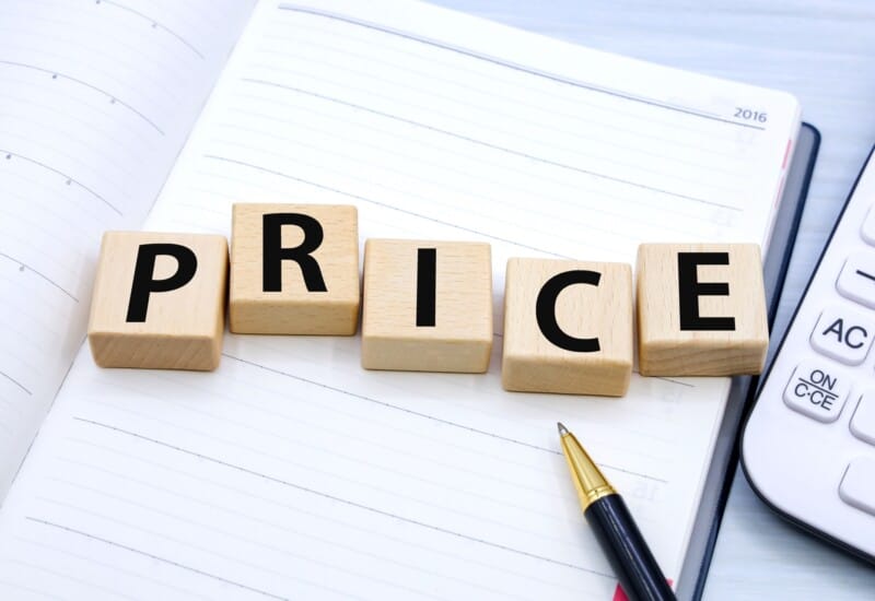 Wooden letter blocks spelling PRICE on an open notebook with a pen nearby and a calculator in view, conveying pricing.