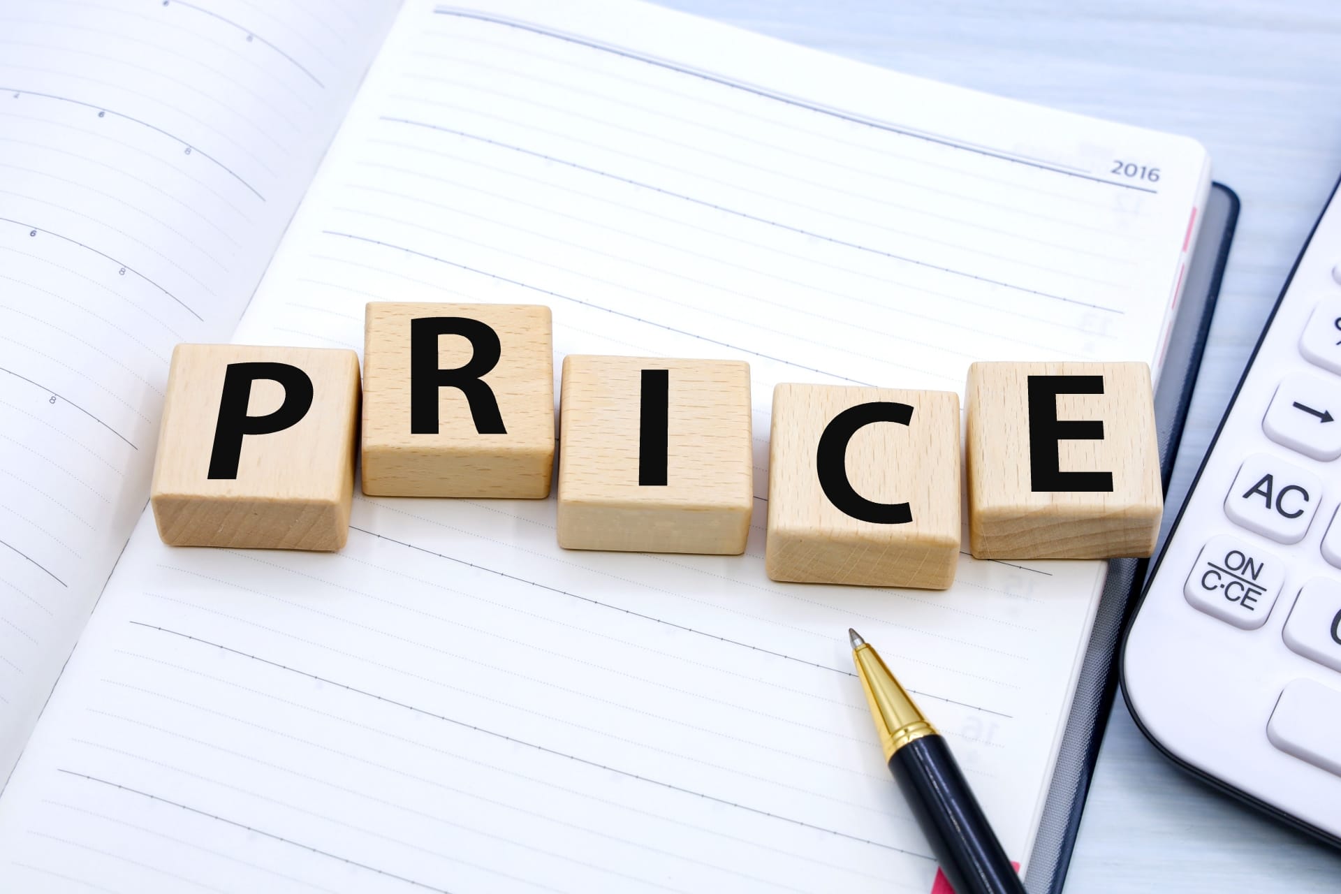 Wooden letter blocks spelling PRICE on an open notebook with a pen nearby and a calculator in view, conveying pricing.