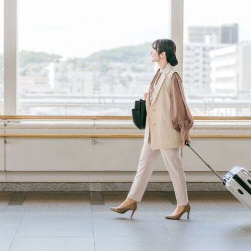 Woman in a beige suit walking through an airport with a rolling suitcase in tow to the right side of the frame.