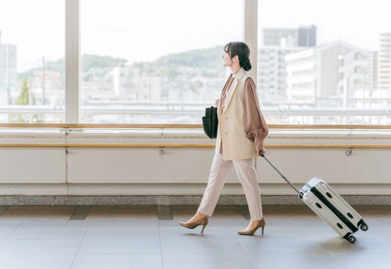 Woman in a beige suit walking through an airport with a rolling suitcase in tow to the right side of the frame.