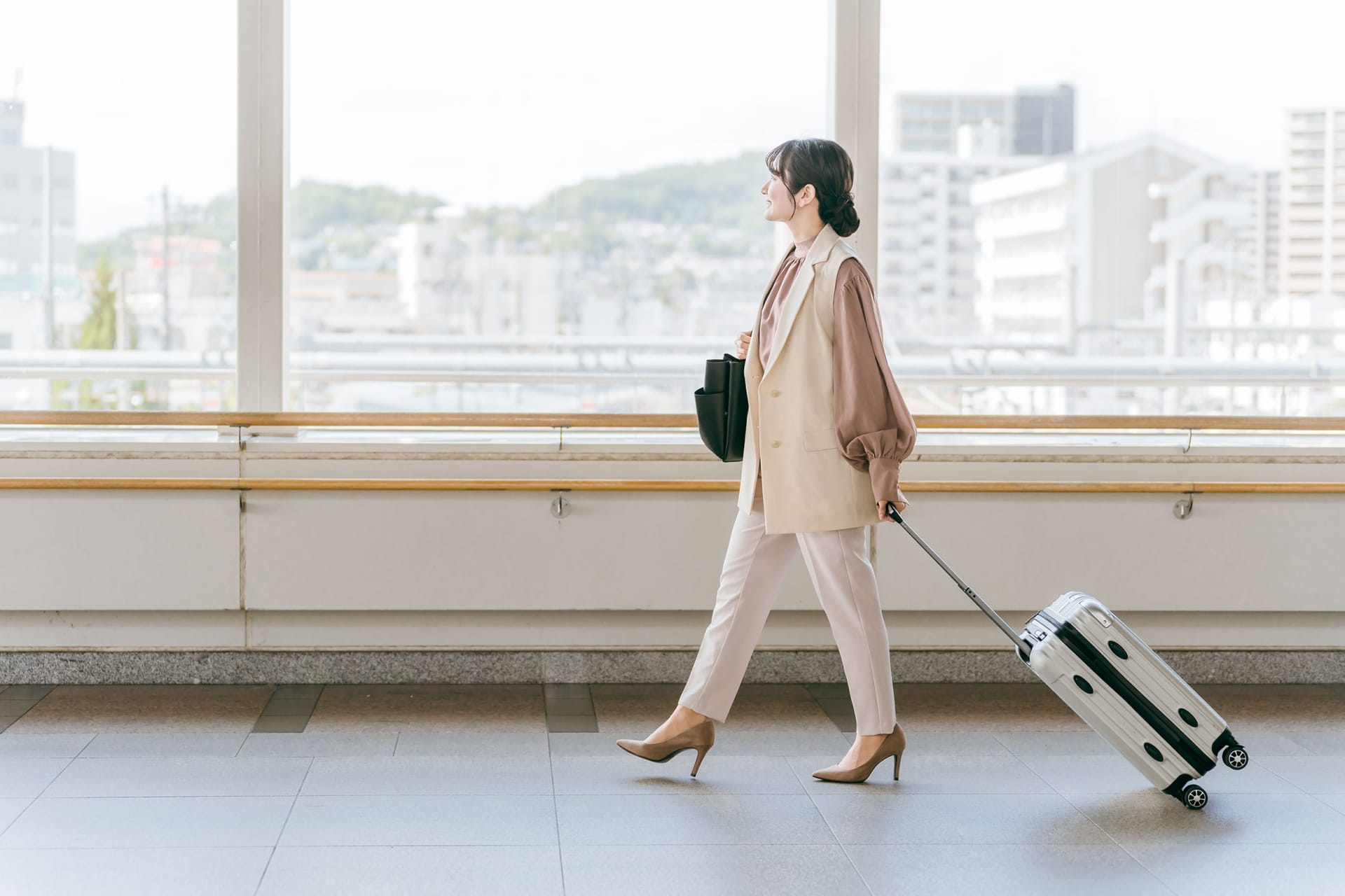 Woman in a beige suit walking through an airport with a rolling suitcase in tow to the right side of the frame.