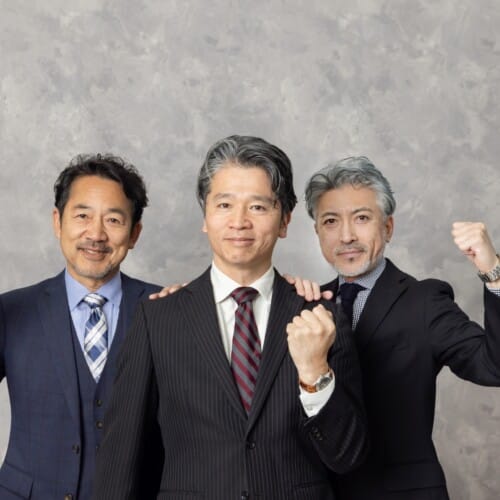 Three businessmen in suits pose together, fists raised in celebration against a gray backdrop, smiling confidently at the camera.