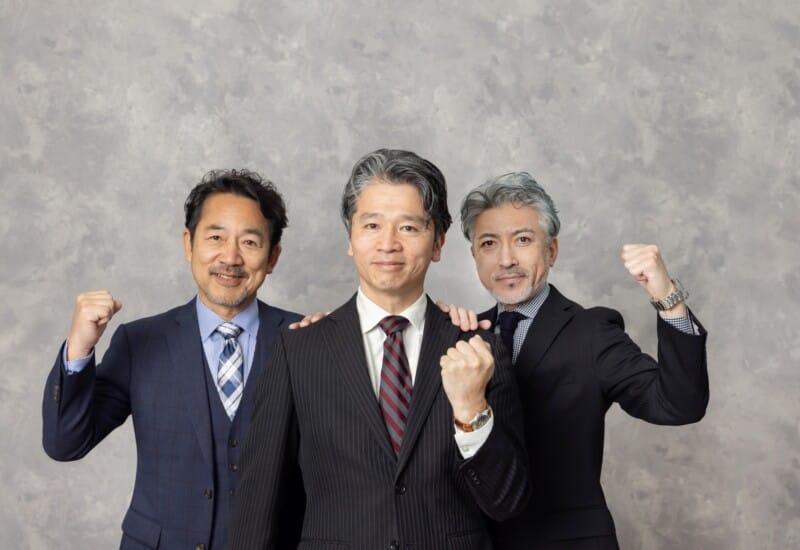 Three businessmen in suits pose together, fists raised in celebration against a gray backdrop, smiling confidently at the camera.