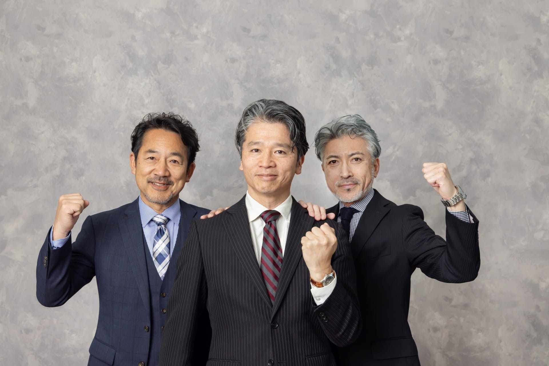Three businessmen in suits pose together, fists raised in celebration against a gray backdrop, smiling confidently at the camera.