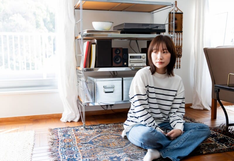 Young woman in a striped sweater sits cross-legged on a patterned rug in a bright living room, holding a smartphone beside a metal shelving unit with stereo equipment.