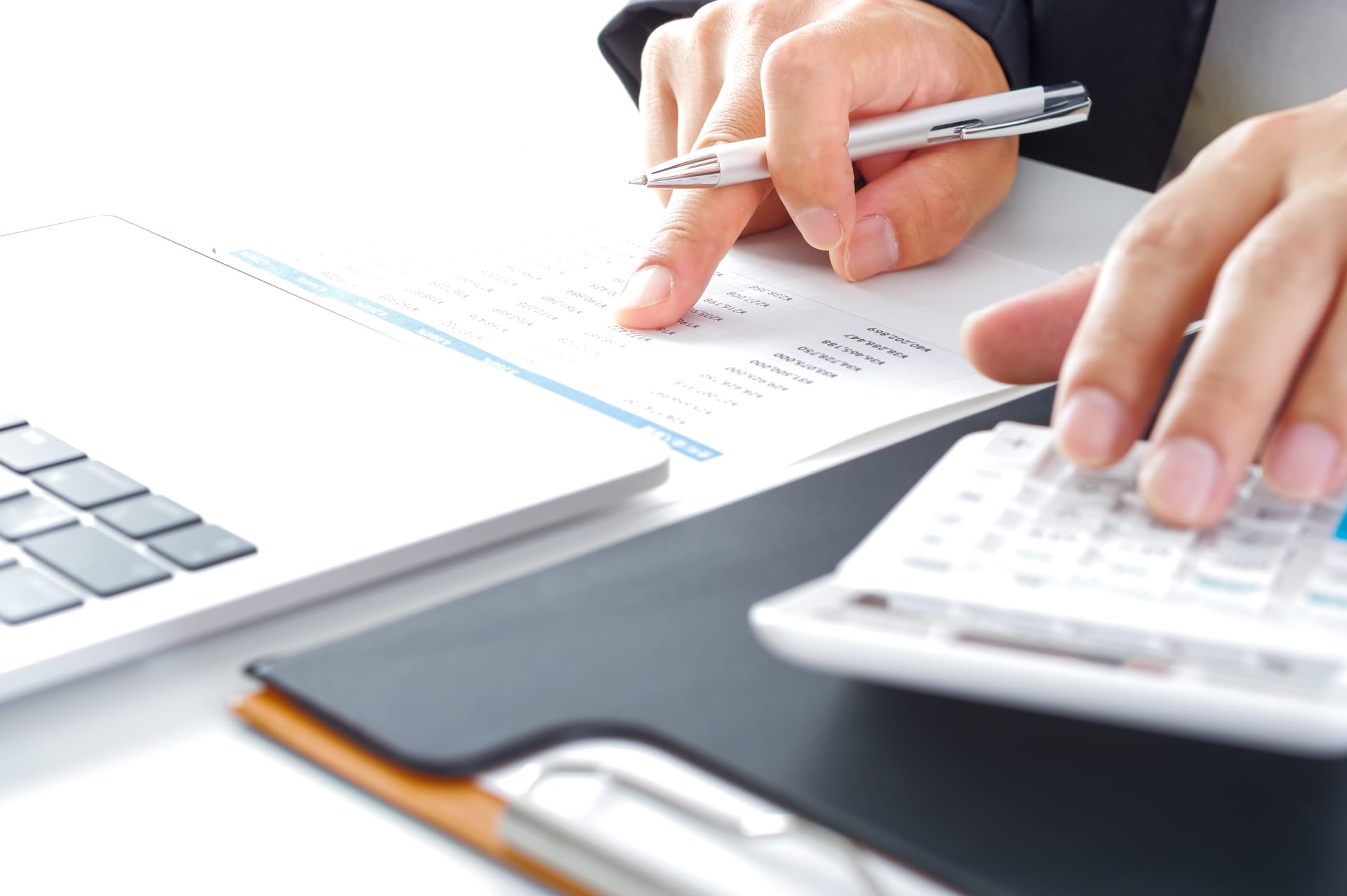 Person writes on a paper document with a silver pen, calculator and laptop nearby on a desk