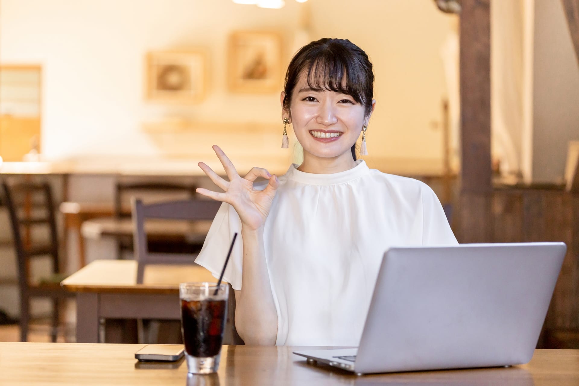 Smiling woman in a café using a laptop, making an OK gesture with her right hand.