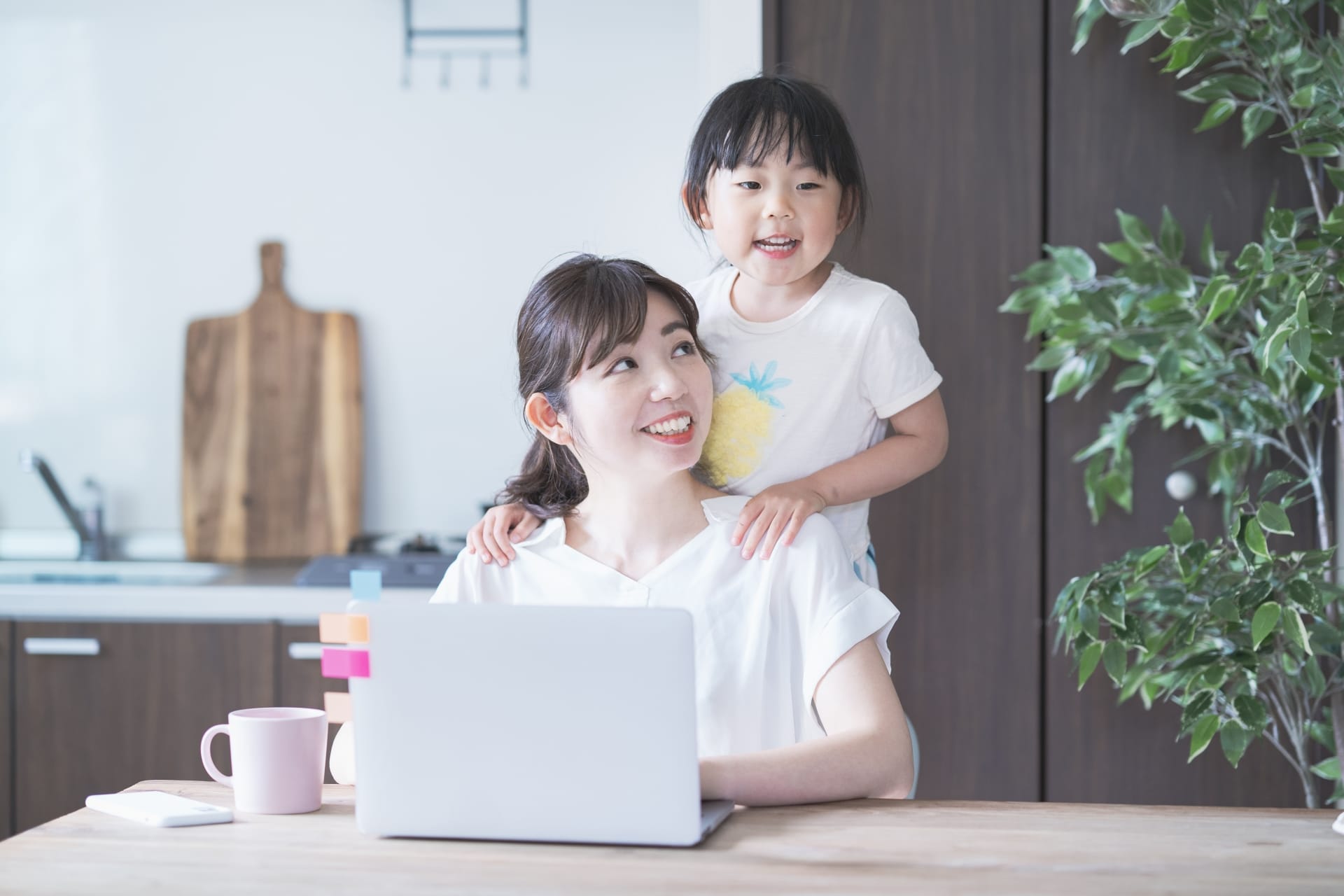 Mother typing on a laptop at a kitchen table with her young daughter perched on her shoulders.