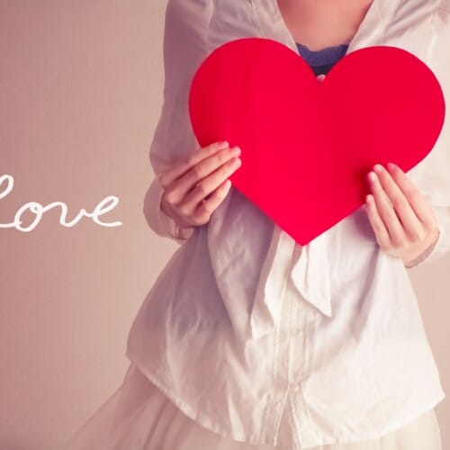 Person in a white outfit holds a large red heart, with the word 'Love' written in white on the left side.