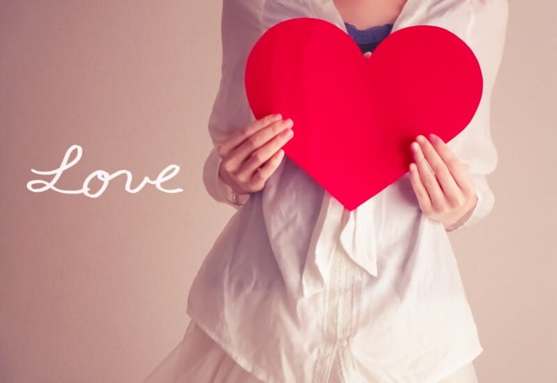 Person in a white outfit holds a large red heart, with the word 'Love' written in white on the left side.