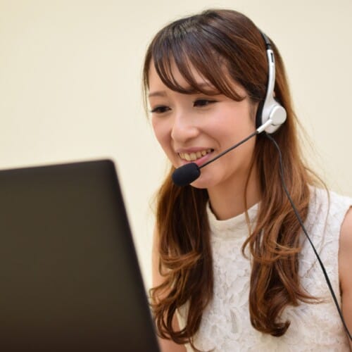 Smiling woman wearing a headset with microphone while looking at a laptop, likely a call center or customer service agent.
