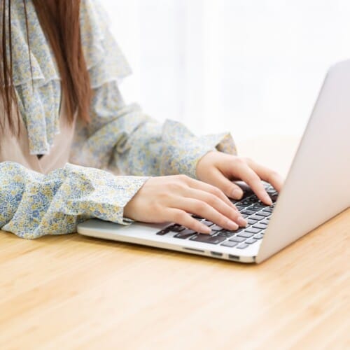 Person typing on a silver laptop at a wooden table, wearing a blue-yellow floral blouse on long sleeves.