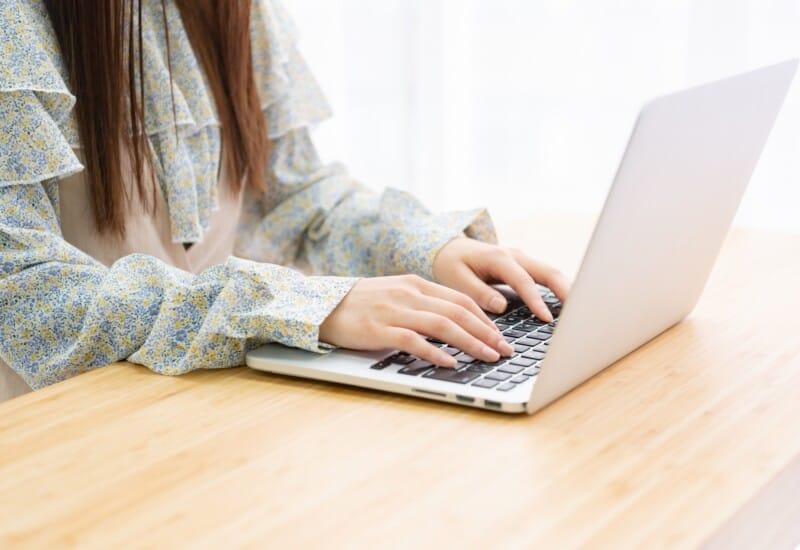 Person typing on a silver laptop at a wooden table, wearing a blue-yellow floral blouse on long sleeves.