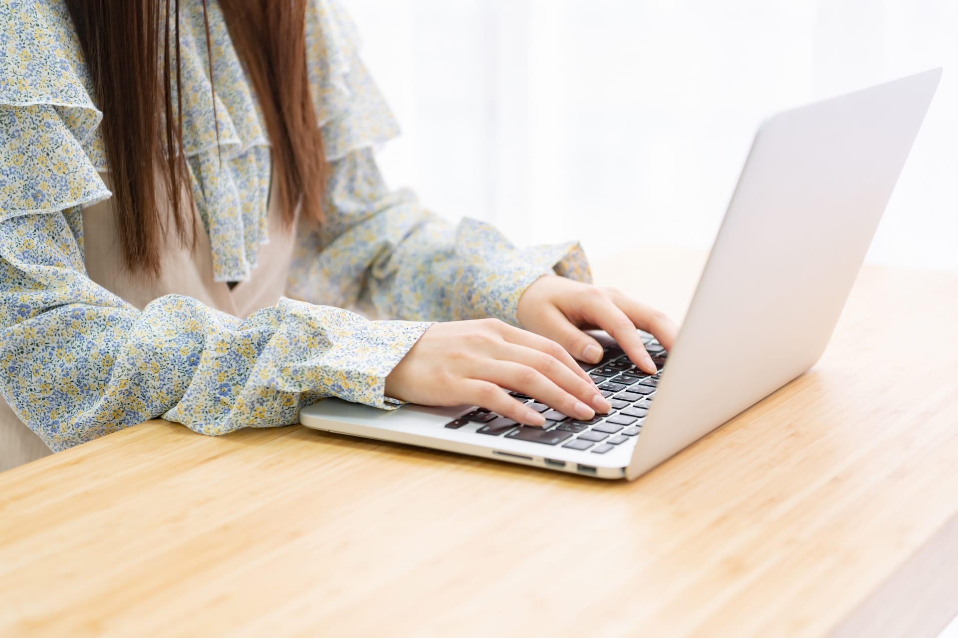 Person typing on a silver laptop at a wooden table, wearing a blue-yellow floral blouse on long sleeves.
