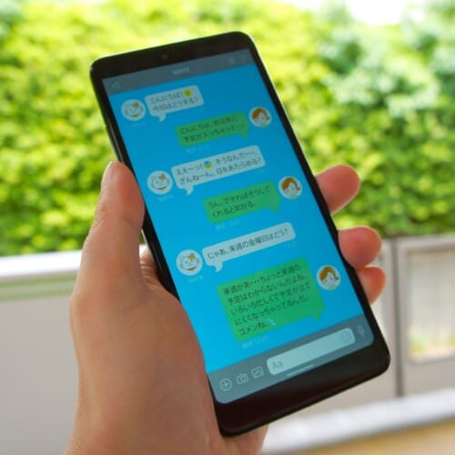 Person holding a smartphone outdoors, screen shows a blue chat app with chat bubbles and avatars (Japanese text).