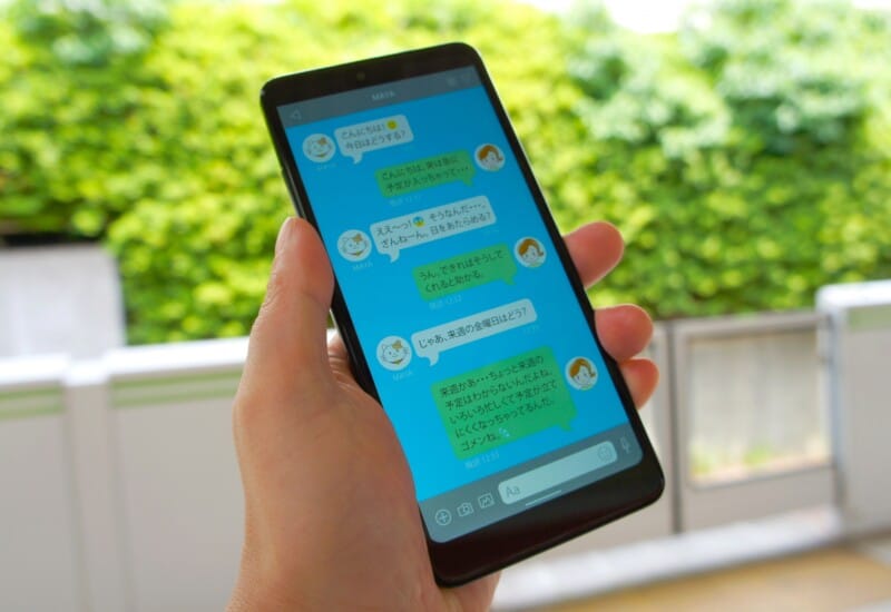 Person holding a smartphone outdoors, screen shows a blue chat app with chat bubbles and avatars (Japanese text).