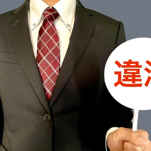 Person in a dark suit and red plaid tie holding a white circular sign with red Japanese characters 違法 (illegal).