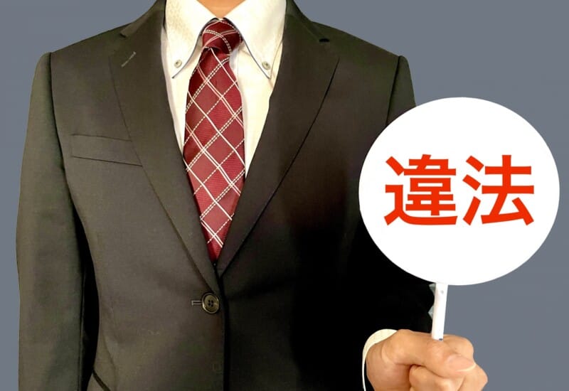 Person in a dark suit and red plaid tie holding a white circular sign with red Japanese characters 違法 (illegal).