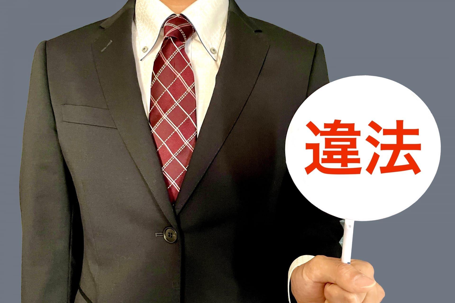 Person in a dark suit and red plaid tie holding a white circular sign with red Japanese characters 違法 (illegal).