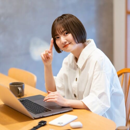 Woman in a white shirt sits at a wooden table with a laptop, smiling and raising her index finger as if making a point.