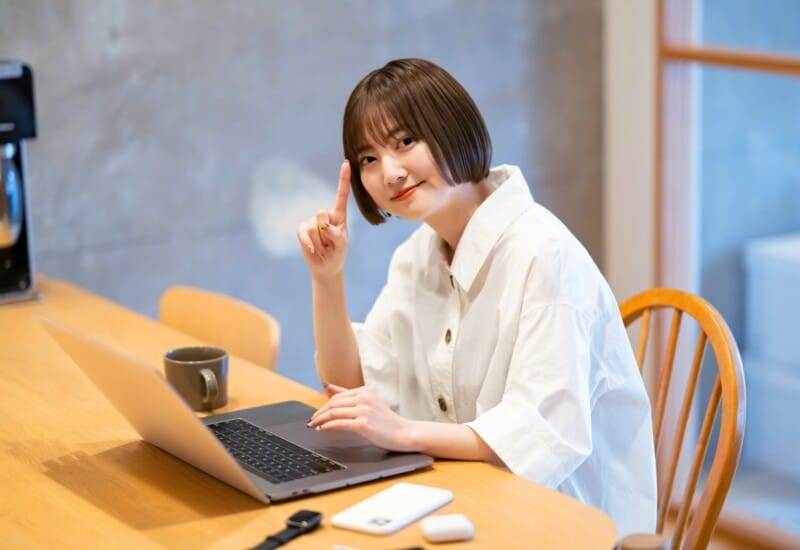 Woman in a white shirt sits at a wooden table with a laptop, smiling and raising her index finger as if making a point.