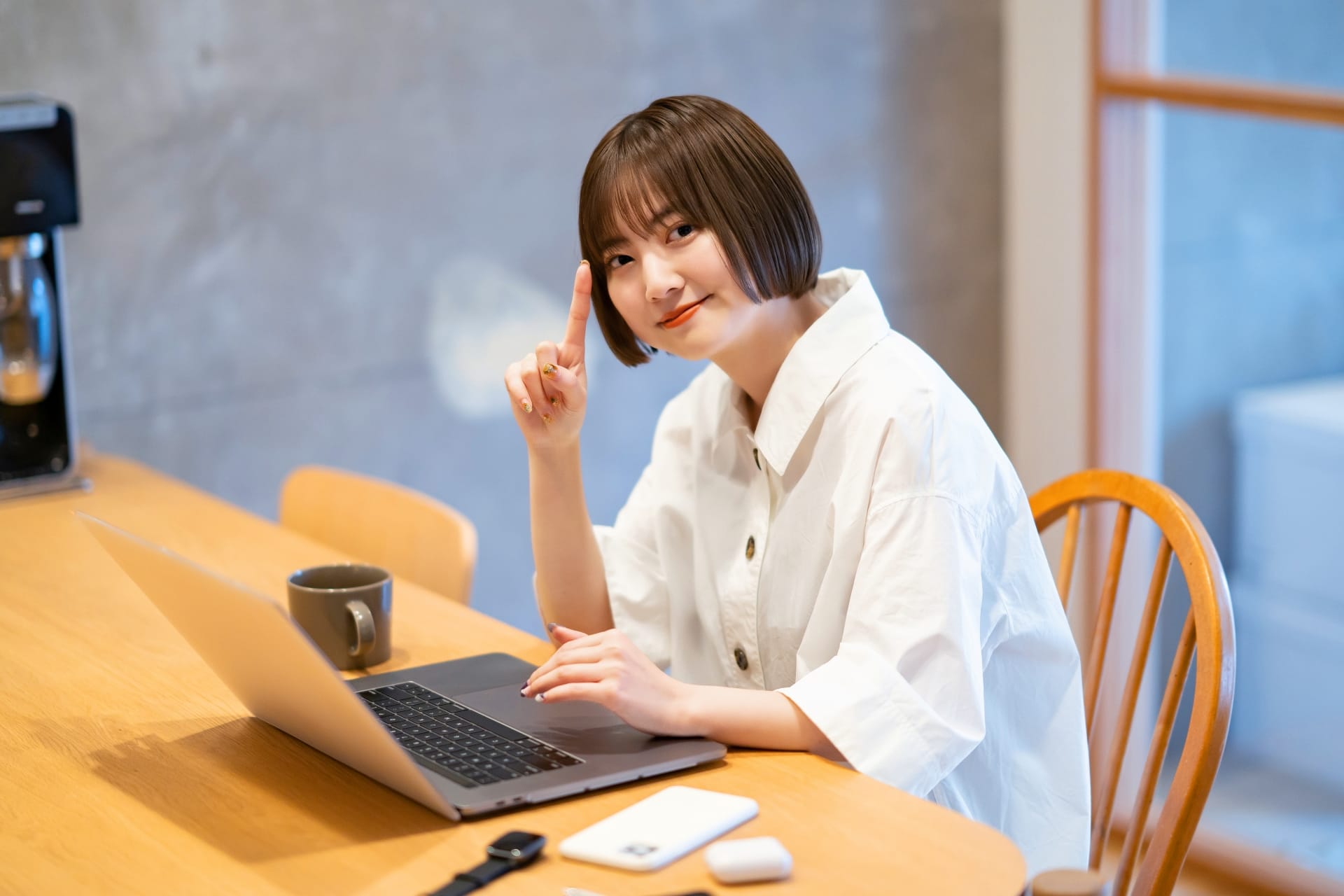 Woman in a white shirt sits at a wooden table with a laptop, smiling and raising her index finger as if making a point.