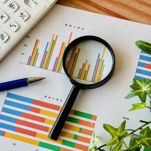 Magnifying glass over colorful bar charts on a desk with a calculator and blue pen nearby.