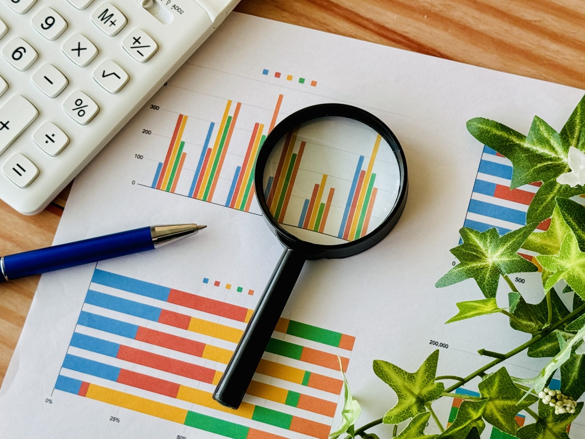Magnifying glass over colorful bar charts on a desk with a calculator and blue pen nearby.
