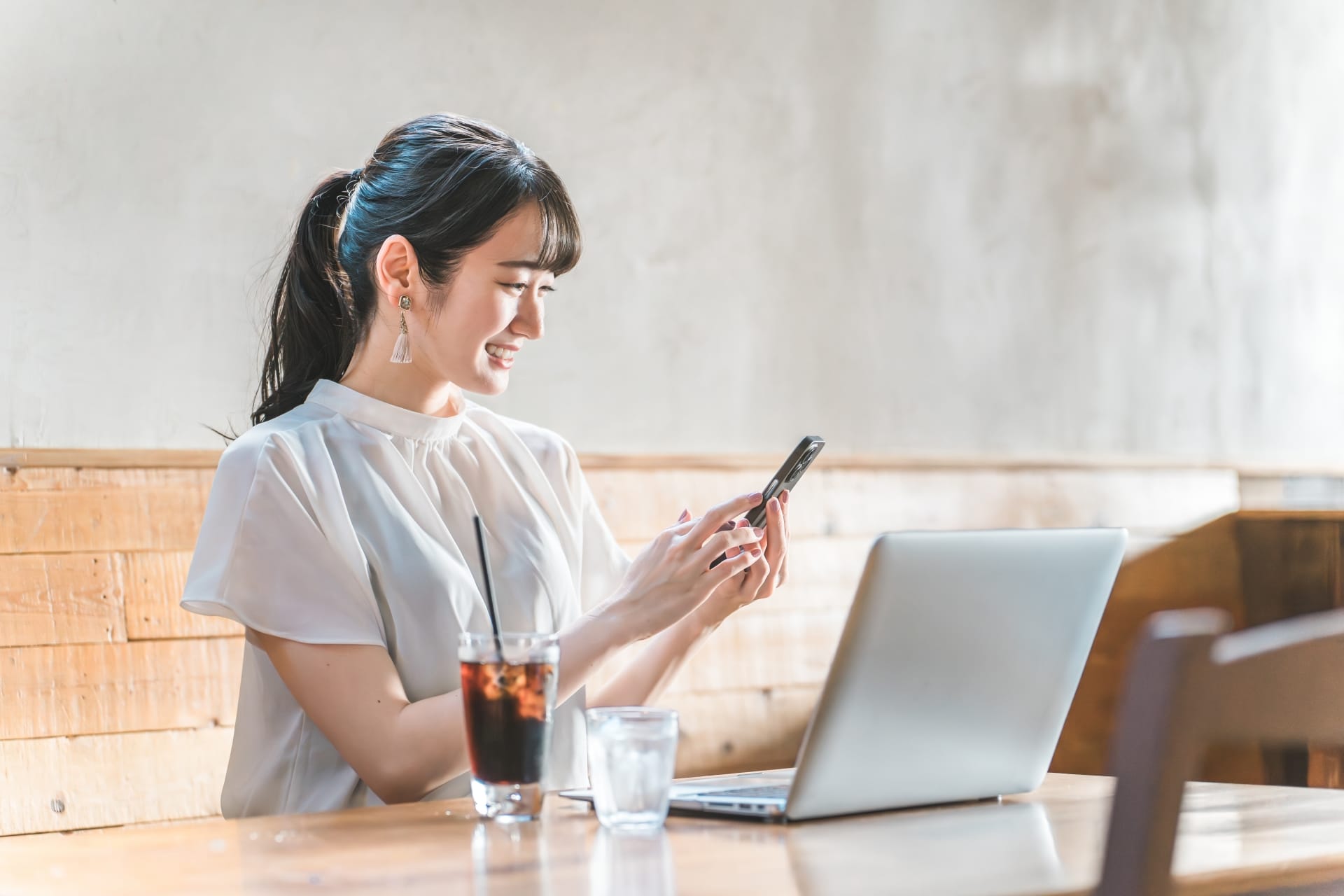 Woman at a cafe table, smiling while looking at her smartphone, with a laptop and iced coffee in view