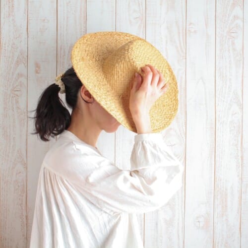 4748197_m - Belle Work Person in a white blouse holds a straw hat up to cover their face against a pale wooden plank backdrop.
