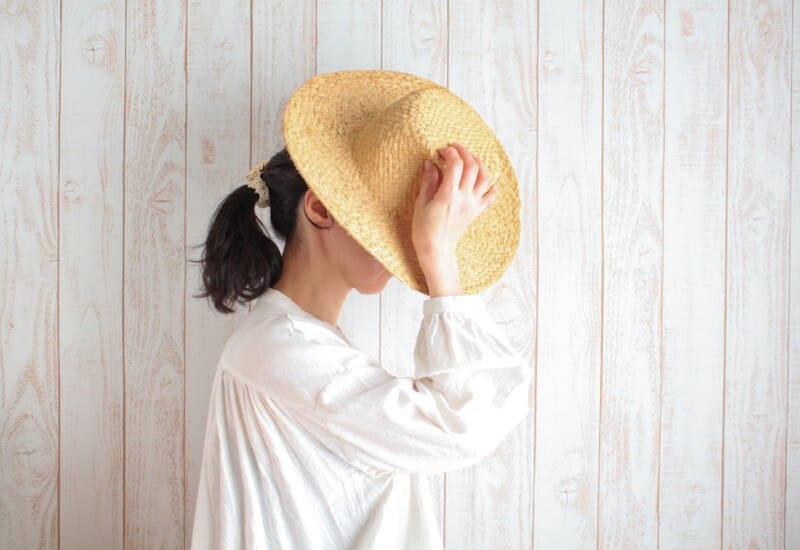 4748197_m - Belle Work Person in a white blouse holds a straw hat up to cover their face against a pale wooden plank backdrop.