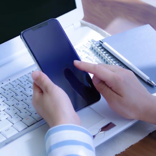 4926757_m - Belle Work Hands hold a smartphone over a white laptop keyboard with a notebook and pen on the desk behind.