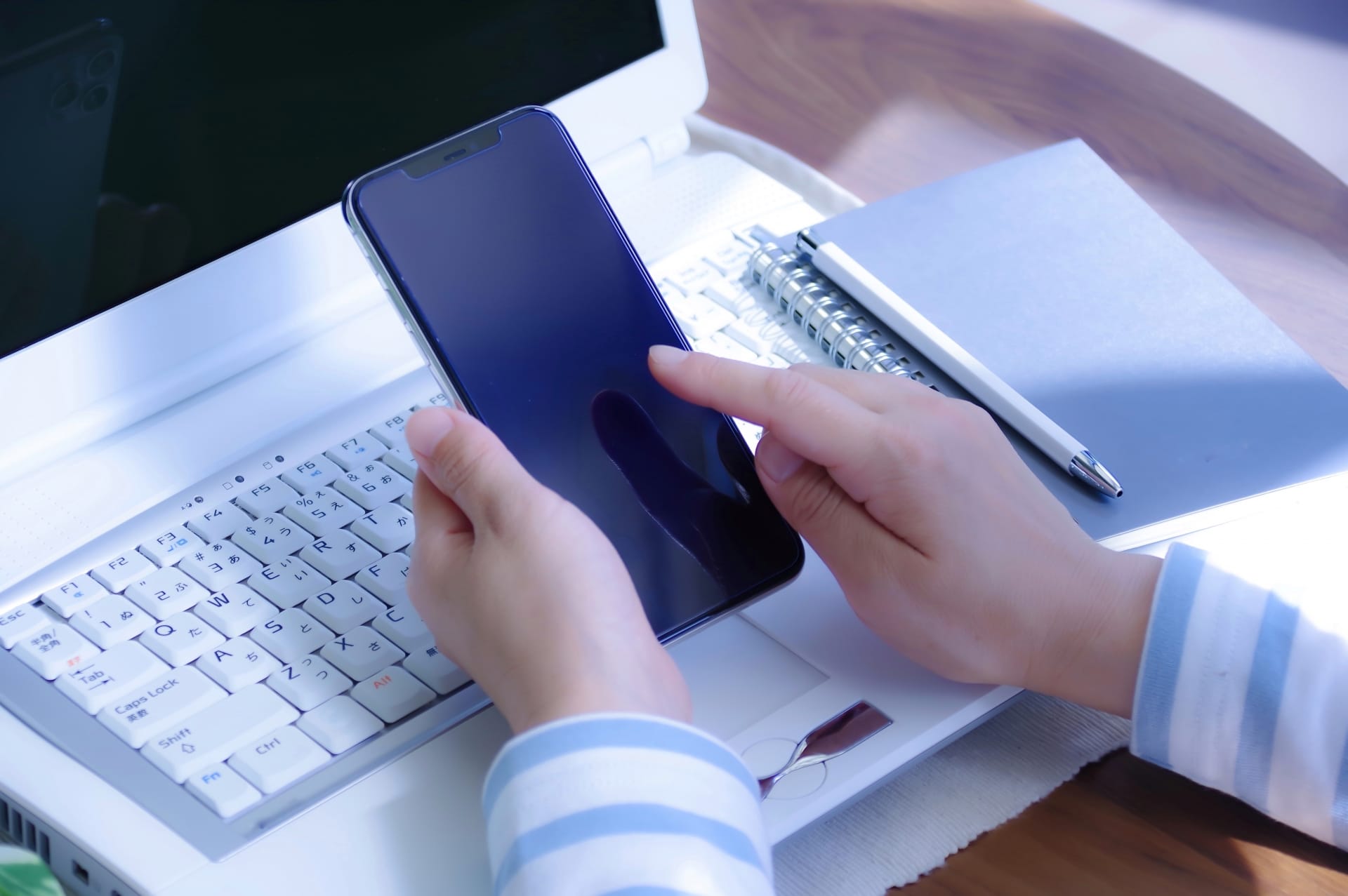 Hands hold a smartphone over a white laptop keyboard with a notebook and pen on the desk behind.